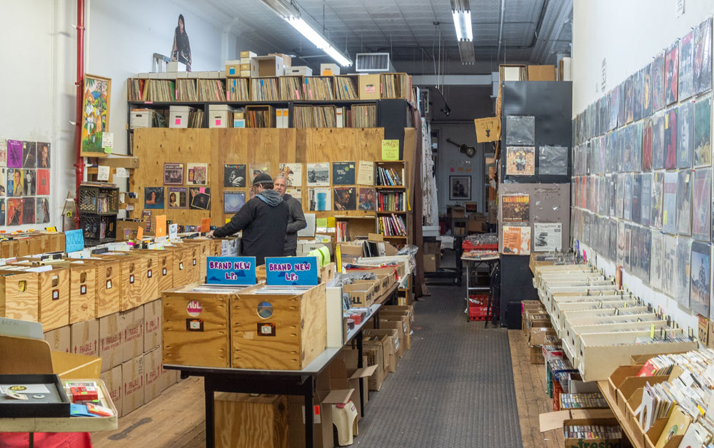 Archive of Contemporary Music in New York City. A man looking through a crate of records in a room filled with them. Vinyl album sleeves cover the walls.