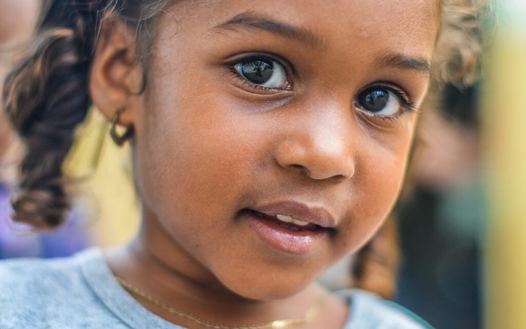 Close up of a young brown-skinned child with curly hair, looking into camera with big, expressive eyes.