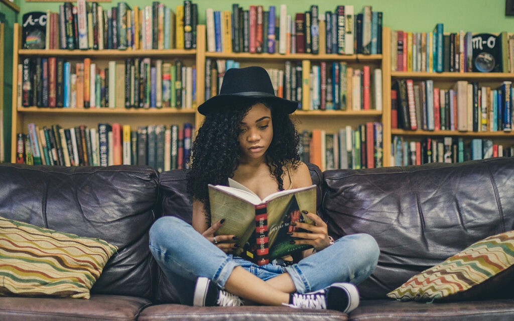 Black girl with a hat sitting on a leather couch reading a book. There is a shelf of books behind her.
