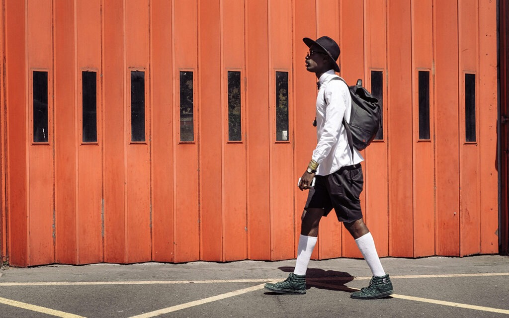 Black man in felt hat, shorts, and high socks walking down the street in front of an orange accordion garage door.