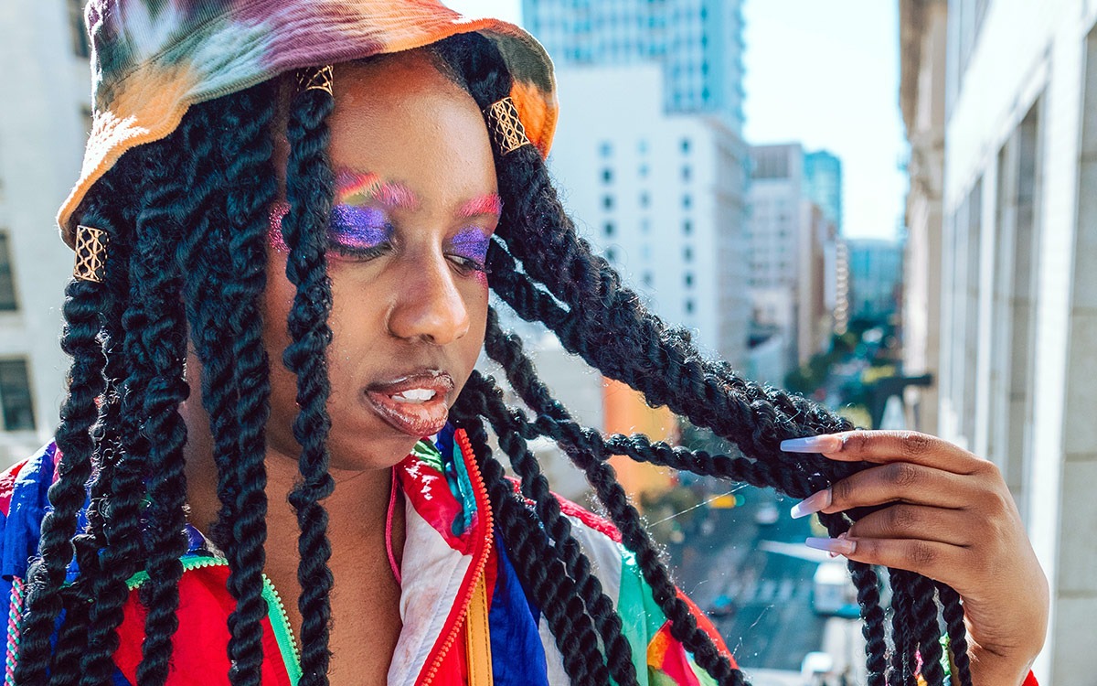 Black woman in colorful jacket, with a tie-die bucket hat, senegalese twists, and long purple nails standing against a city-scape. She is wearing rainbow makeup.