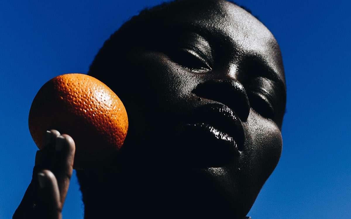Close up photo of dark skinned Black woman against a blue sky. She is holding an orange to her face.