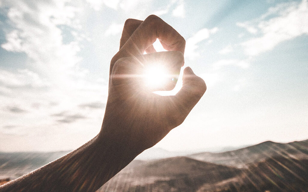 A person making a hole with their fingers, centering the sun in the hole. The sun's rays are shining through the fingers. There are mountains in the background.
