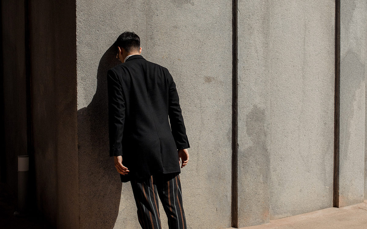 Asian man in black suit jacket and striped pants, facing towards a wall, leaning his head against the wall.