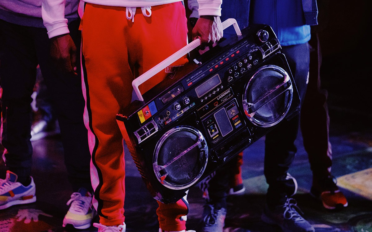 An image of four men, from the waist down in athletic wear. The man in the front is wearing red sweatpants and holding a boom box.