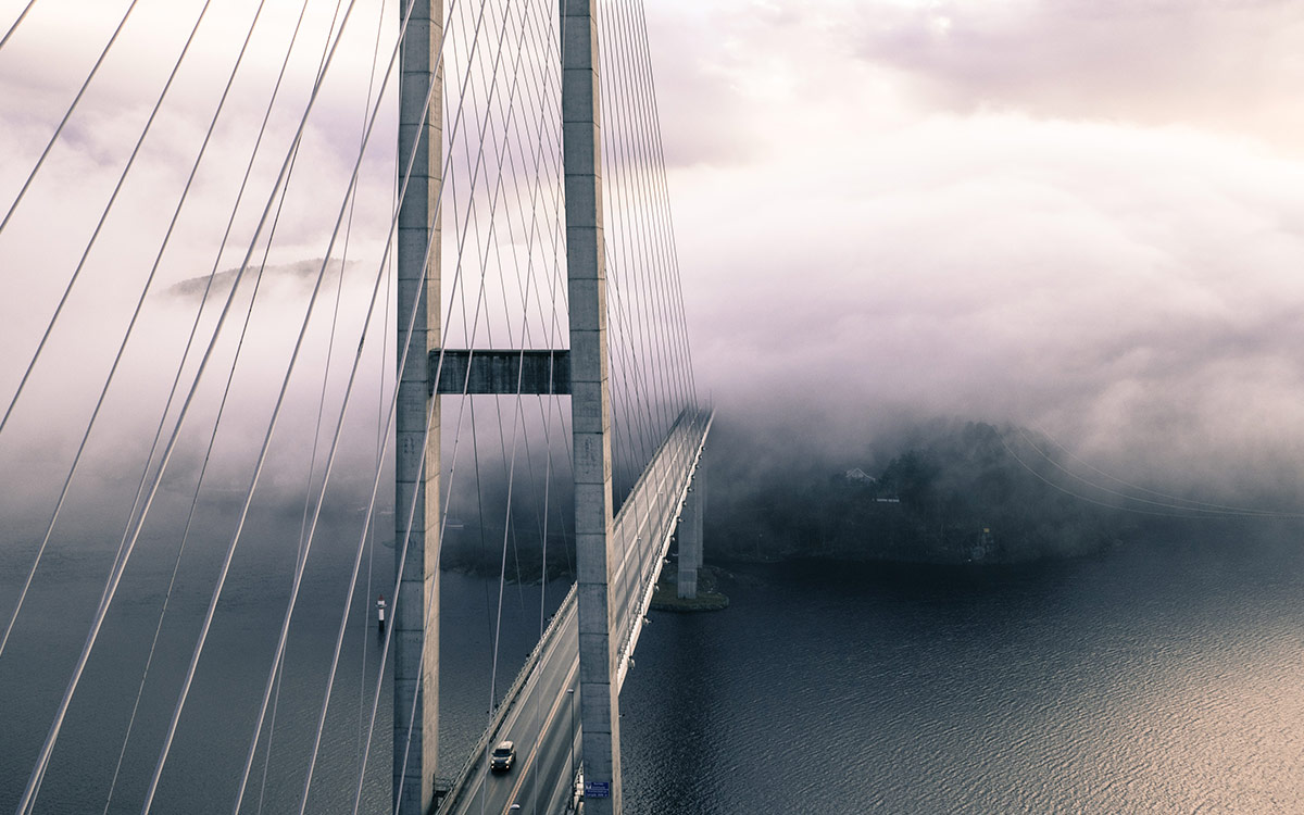 A suspension bridge over a body of water, that leads to a cloud-covered town