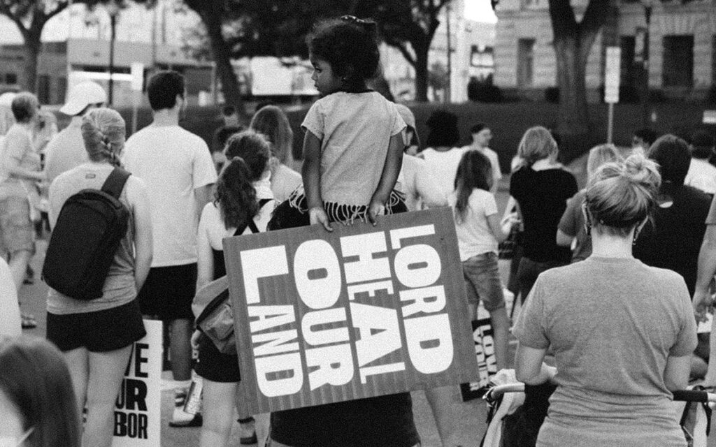 a black and white photo of a demonstration, with a child sitting on a adults shoulders, holding a sign that reads, "Lord, Heal Our Land"