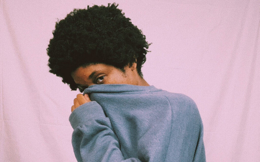 A Black woman with an afro, standing against a pink wall. She is covering most of her face with her sweatshirt.