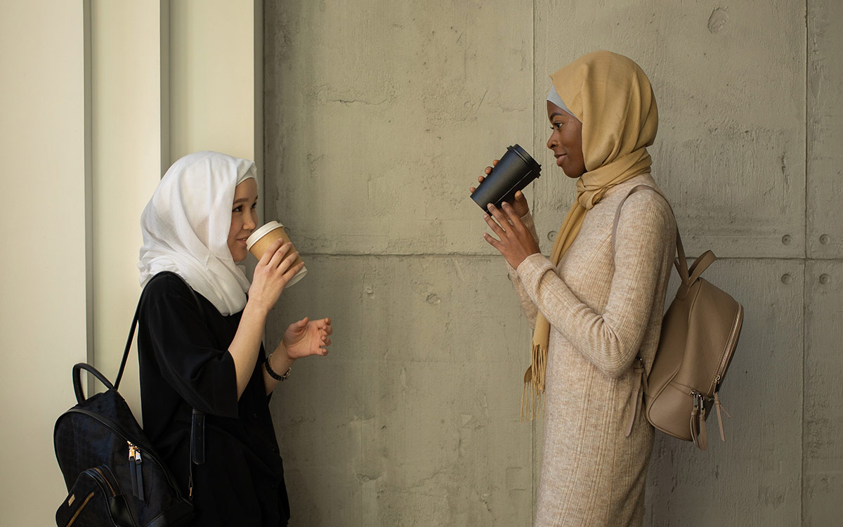 Two women–one Asian and one Black– wearing hijabs and leaning against a wall talking. They are both holding coffee cups.