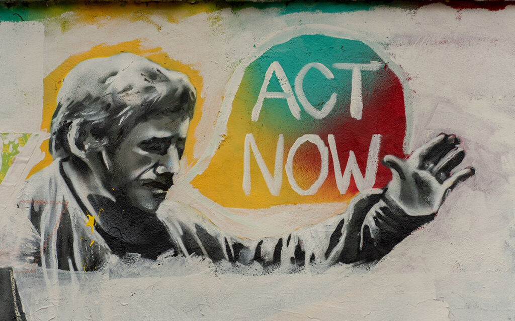 A graffiti piece of a mustachioed man holding up his hand, with a multicolored speech bubble that says "act now"