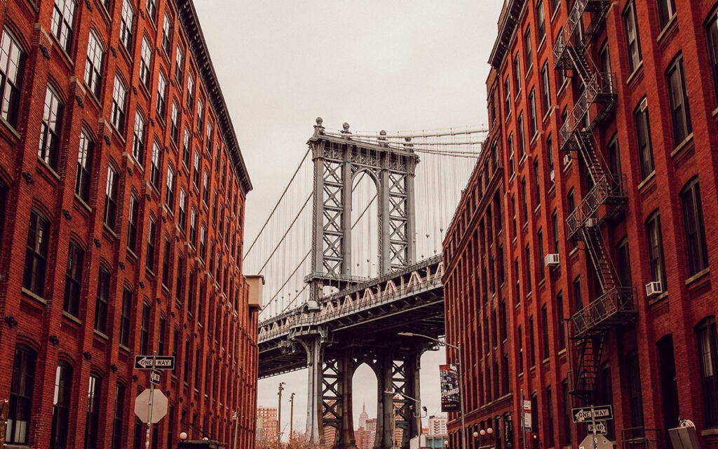 Photograph of the Brooklyn bridge peeking through two brownstones on either side of the street.