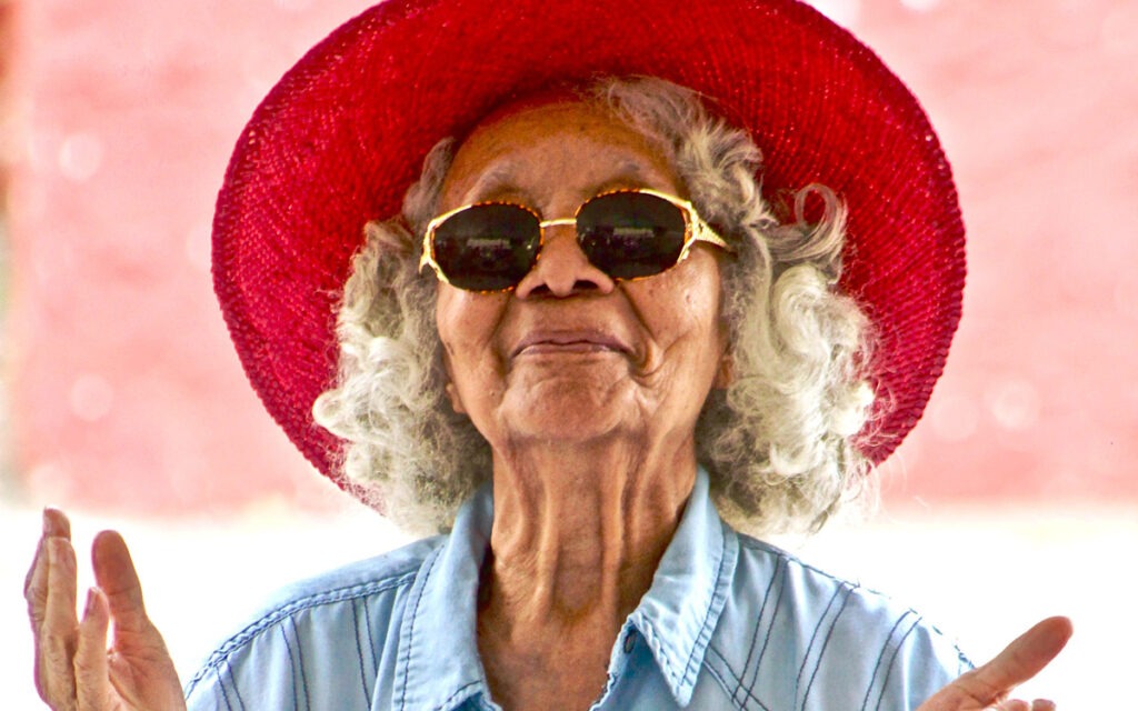 An Asian elder wearing a red, wide-brimmed hat and sunglasses. She is holding her hands up.