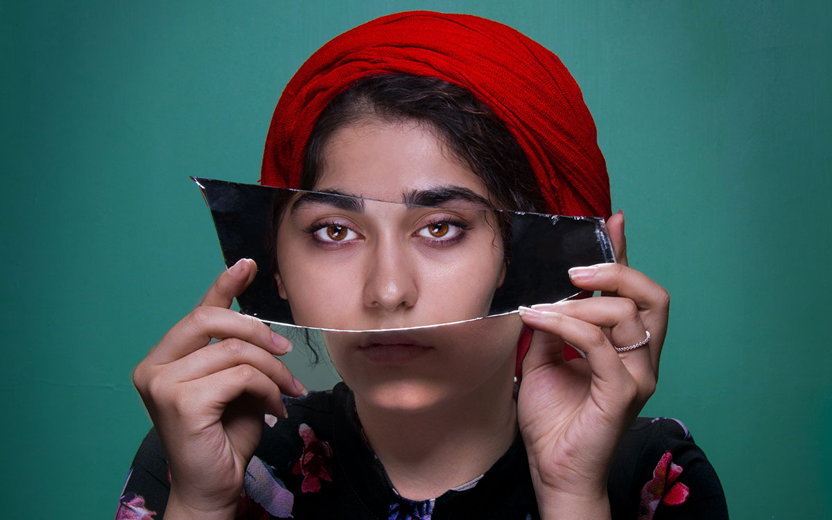 Dusky woman with curly hair wearing a red turban and holding a broken piece of mirror in front of her eyes. There is another woman in the mirror, aligned with the holder.