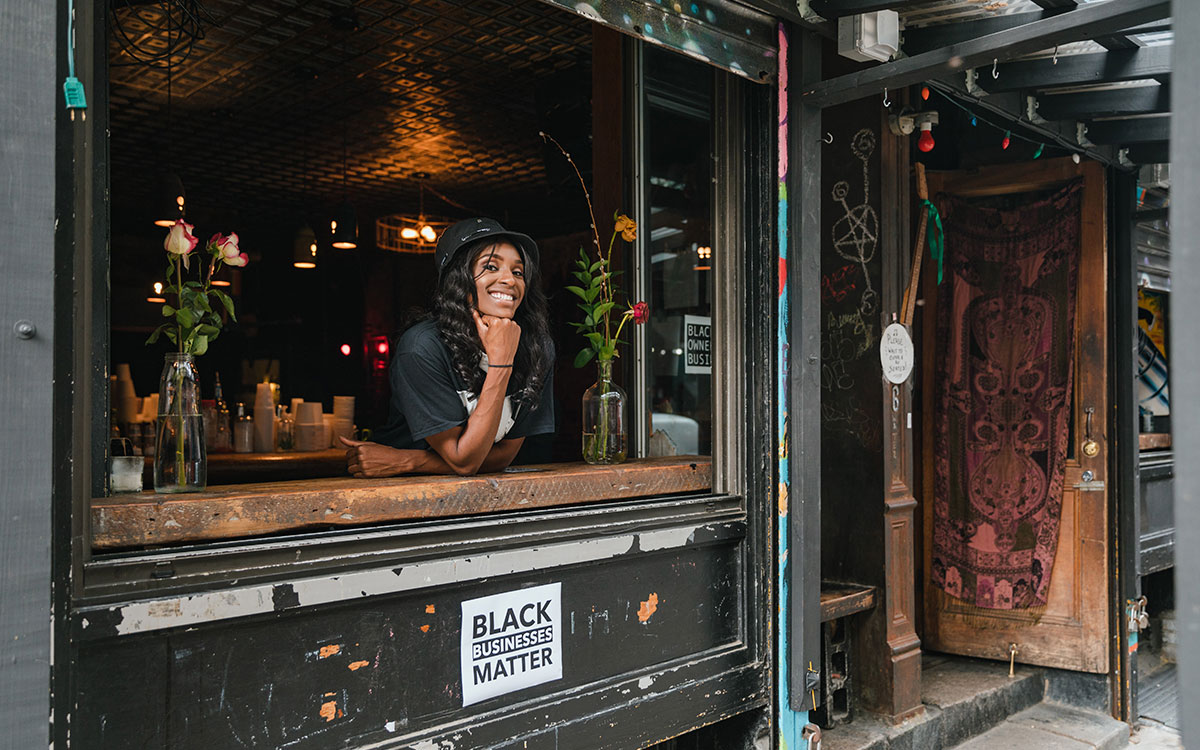 A black woman with a bucket hat smiling and resting on a coffee shop window counter. There is a sign below the counter that reads, "Black Businesses Matter"
