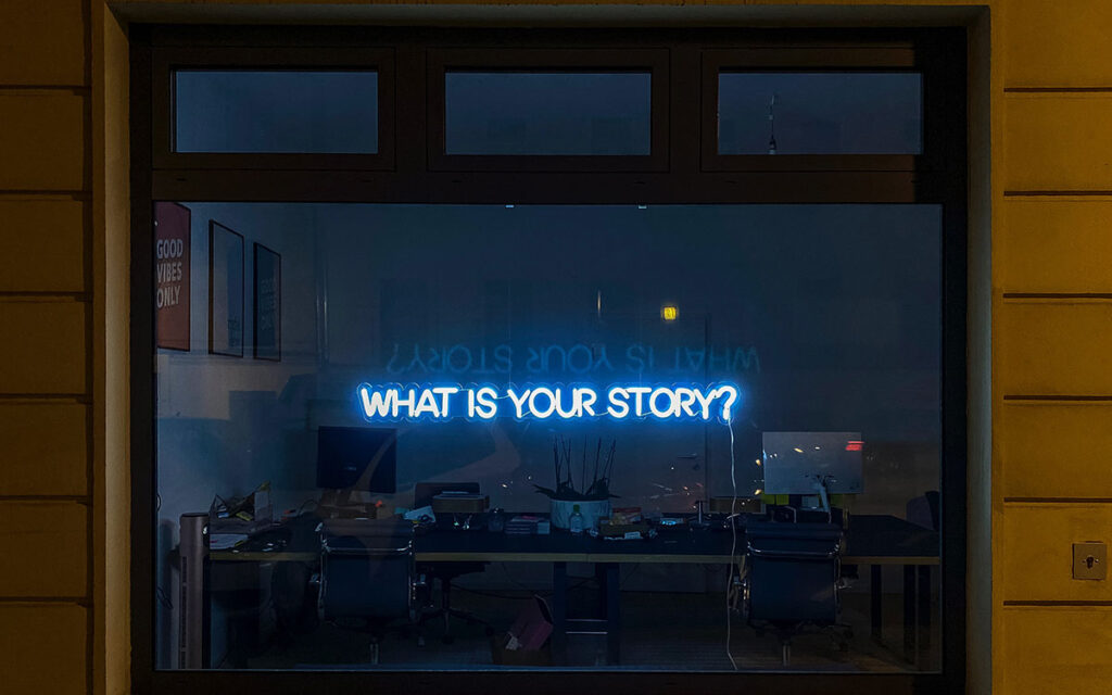 A blue neon sign reading "What is Your Story?" The sign sits in an office window with the lights turned off.