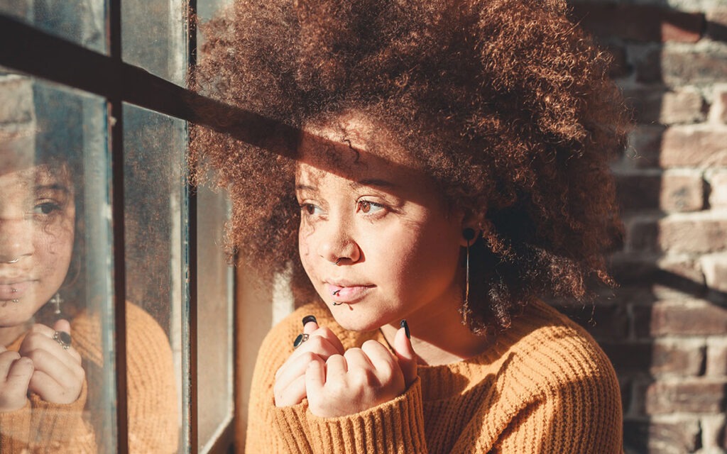 Black woman with auburn afro, looking out the window and holding her hands together on her chest