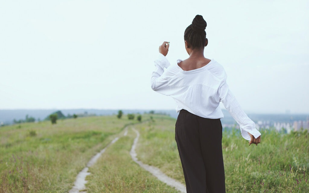 A Black woman with braids sauntering down a grass-covered path. She wears a white shirt and a black, long skirt. Her back is turned to the camera.