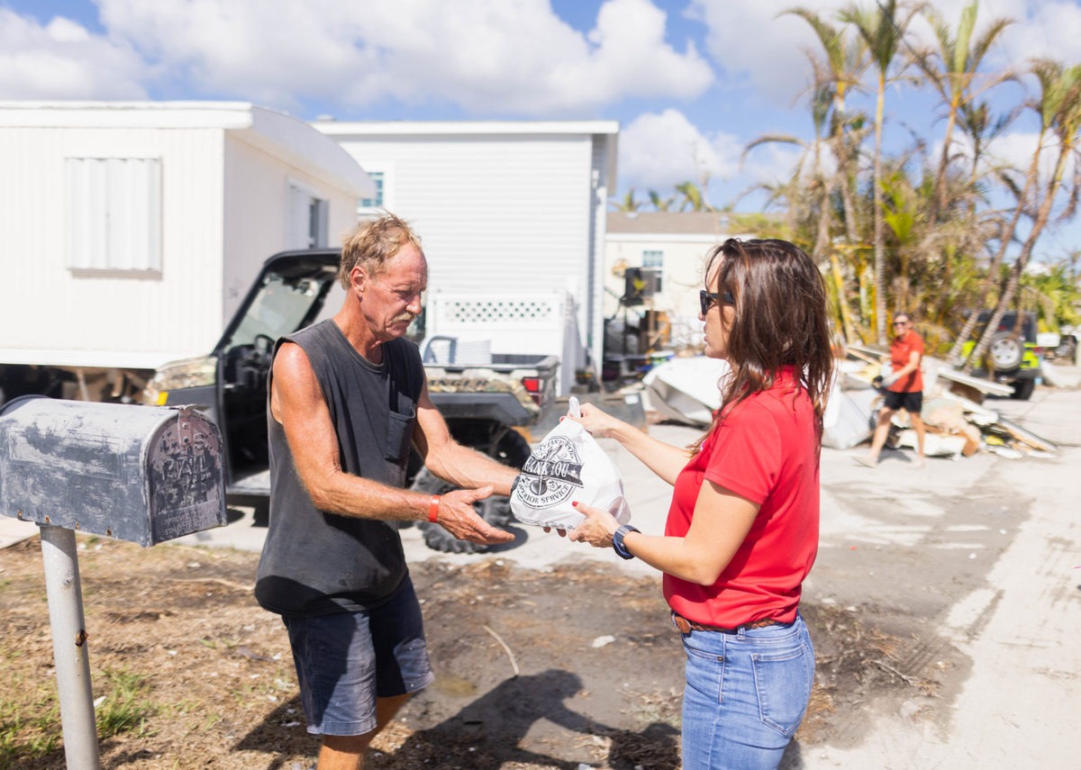 Woman with brown hair handing a bag of food to the blonde man with a mustache. They are standing in a mobile home park.