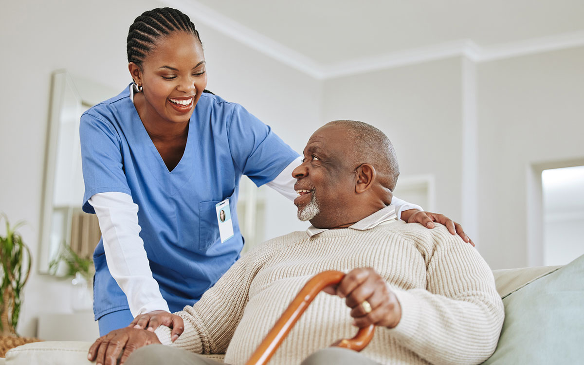 Black nurse in blue scrubs attending to an elderly Black man with a cane. They are both smiling.
