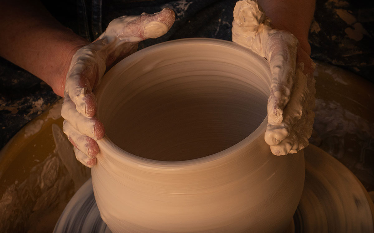 A pair of hands creating a large pot of of clay, on a pottery spinning wheel