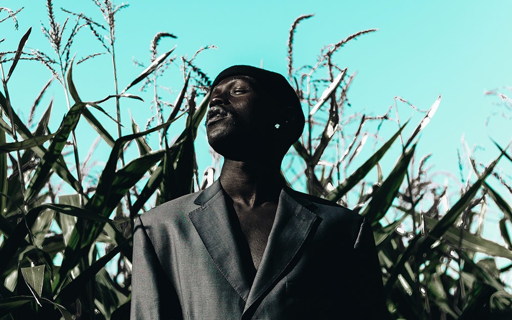 Dark-skinned black man wearing a grey suit jacket standing in a field of wheat. His face is turned to the sun, the sky is clear.