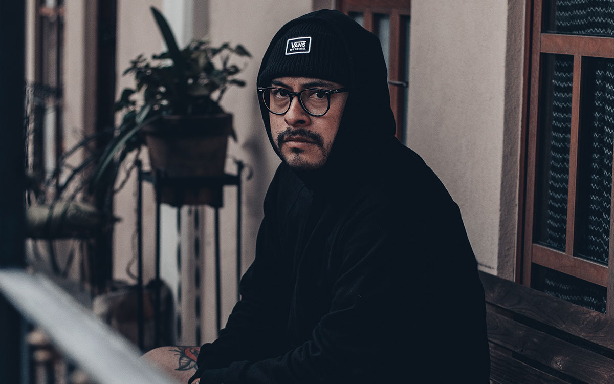 A hispanic man with tattoos and a black hoodie, wearing glasses and sitting on his porch. He is looking into the camera.