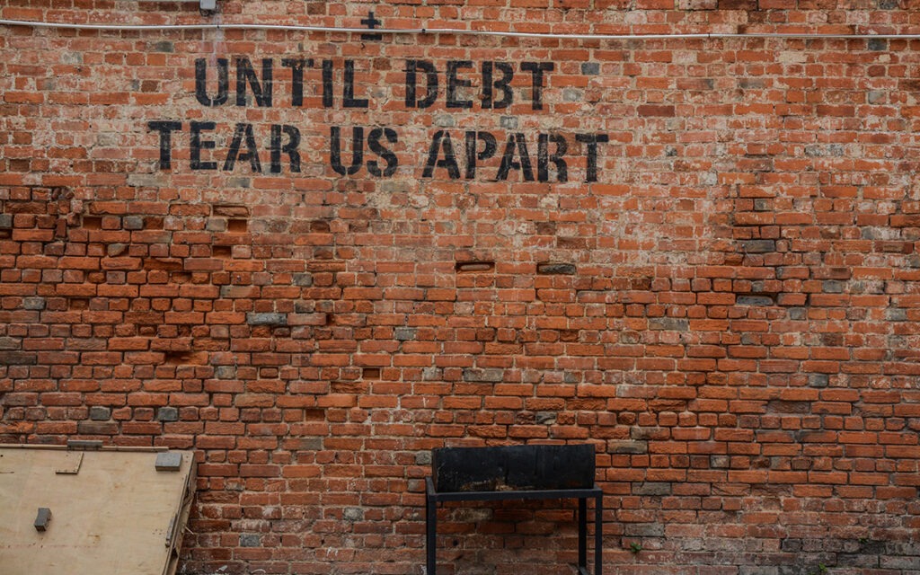 A brick wall in an alley with spray-painted words that read, “Until Debt Tear Us Apart”