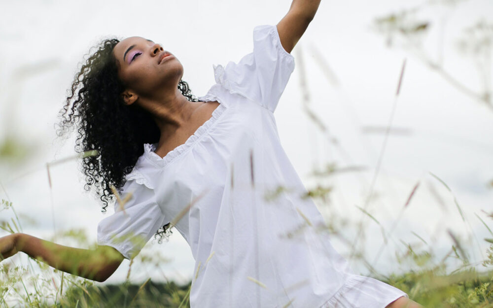 a Black woman with curly hair dances in a field of tall, green grasses.