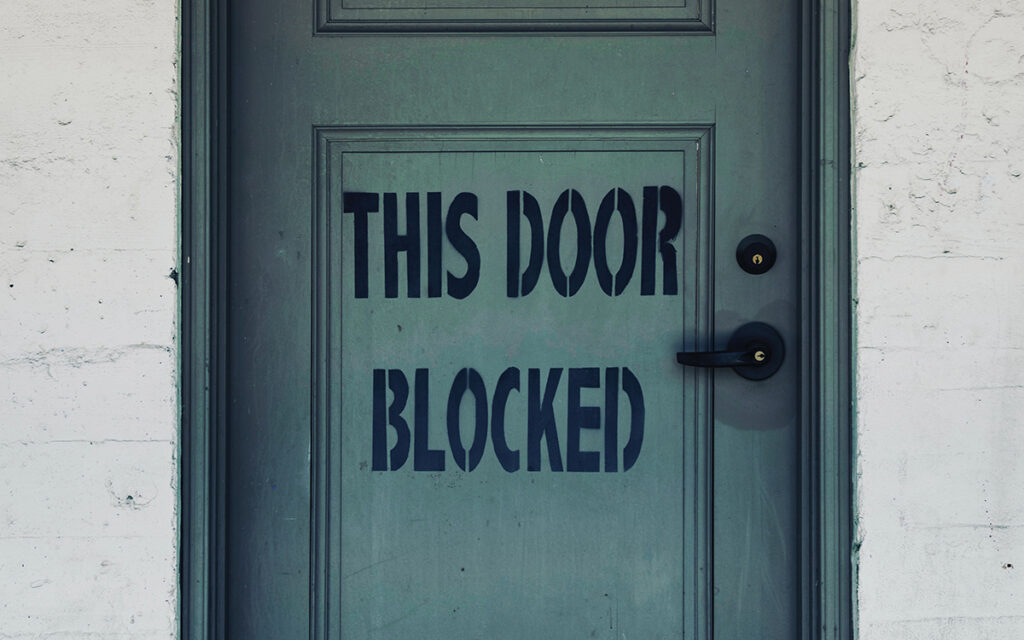 Photograph of blue door with the words, “This Door Blocked” painted on it