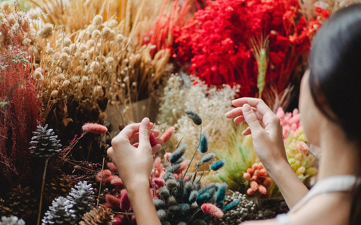alt: A dark-haired woman faces away from the camera, creating an arrangement of colorful dried flowers.