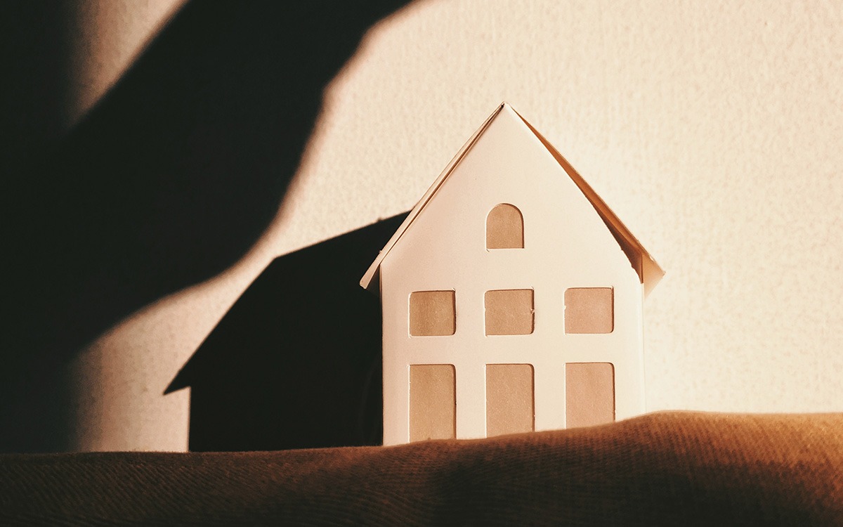 A house made of paper, sitting on a cloth surface. A shadow of a hand looms over the paper structure.