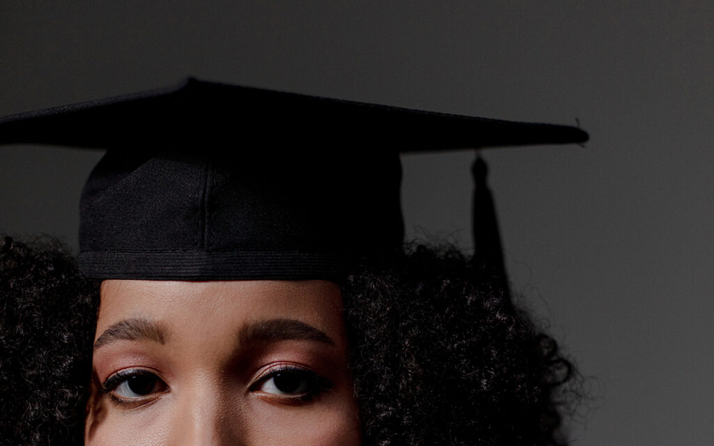 The cropped face of a Black college student with curly hair, wearing a graduation stole. Her expression is serious.