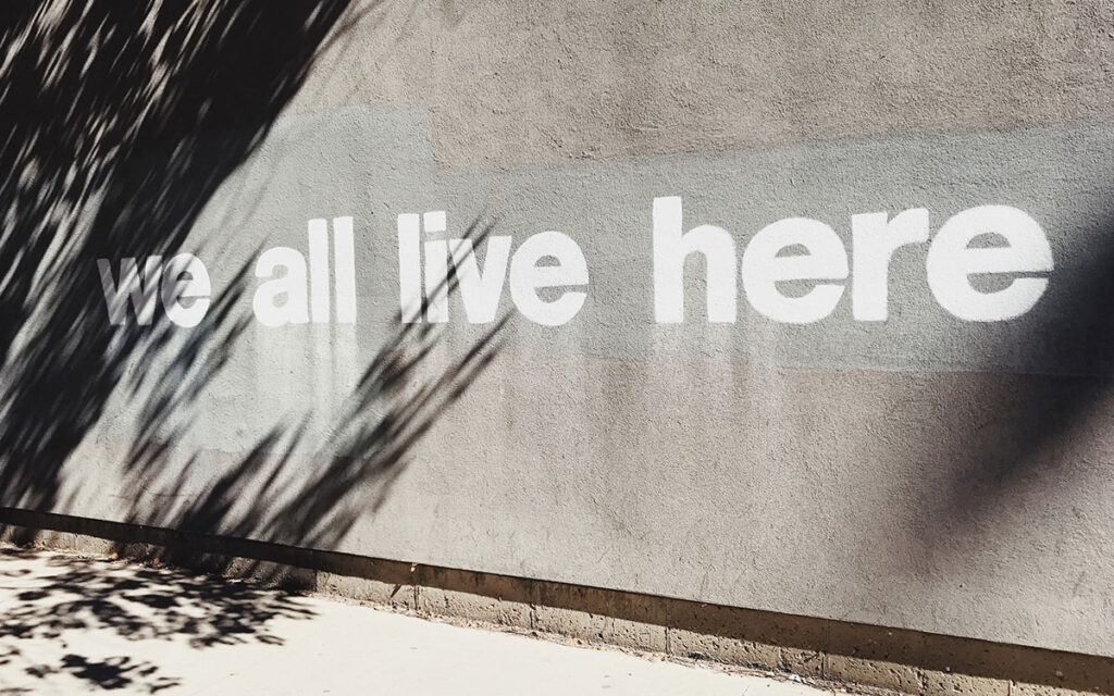 Am outdoor concrete wall with the words “we all live here” painted in white