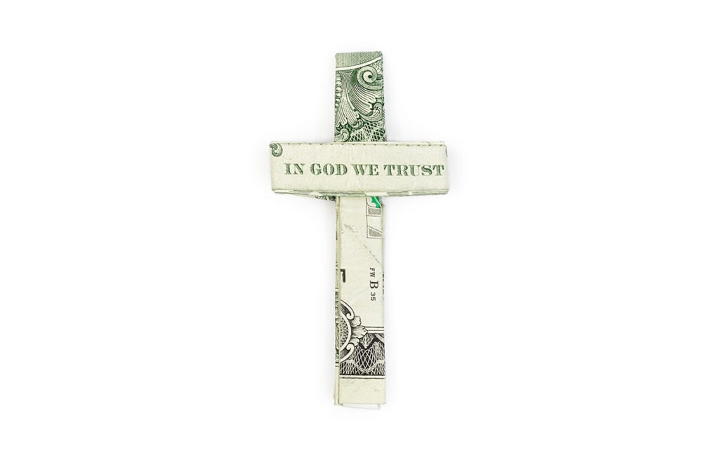A dollar bill folded into the shape of a cross. The horizontal bar shows the words, “In God We Trust”