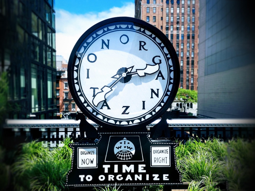 A clock with the words, “Time to organize” painted at the base. Instead of numbers on the clock face, there are letters spelling “Organization”.