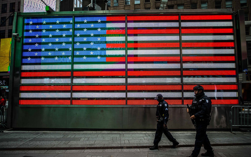 Two armed police officers walking in front of a large lit up American Flag on the side of a building.