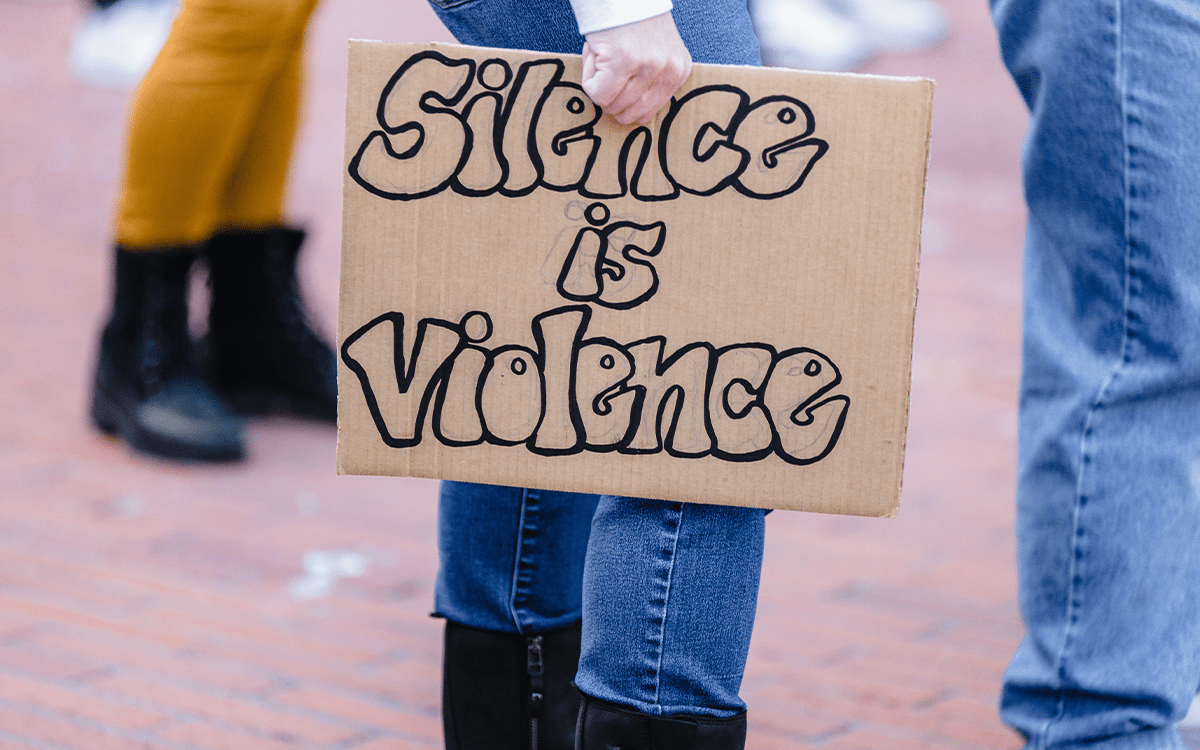 alt: A person holding a cardboard sign with the words “Silence is Violence” written in bubble letters