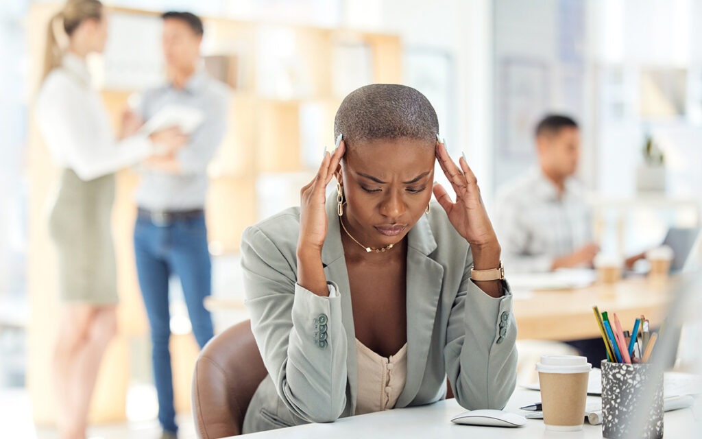 A Black woman, sitting at an office desk with her fingers at her temples in exasperation.