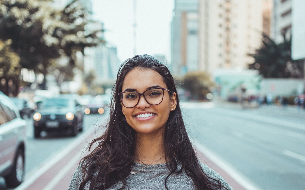A brown-skinned Latinx woman with glasses smiling at the camera. She is standing on a walking path in a street.