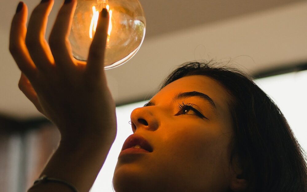 A young, brunette woman holding a large, dimly-lit bulb in the palm of her hand. Her face is washed in a soft glow