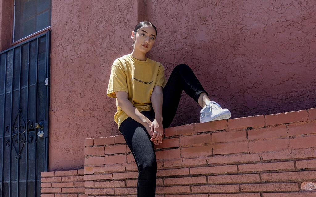 A Latinx woman wearing a yellow shirt and sitting on a brick ledge. She is looking into the camera with a serious expression