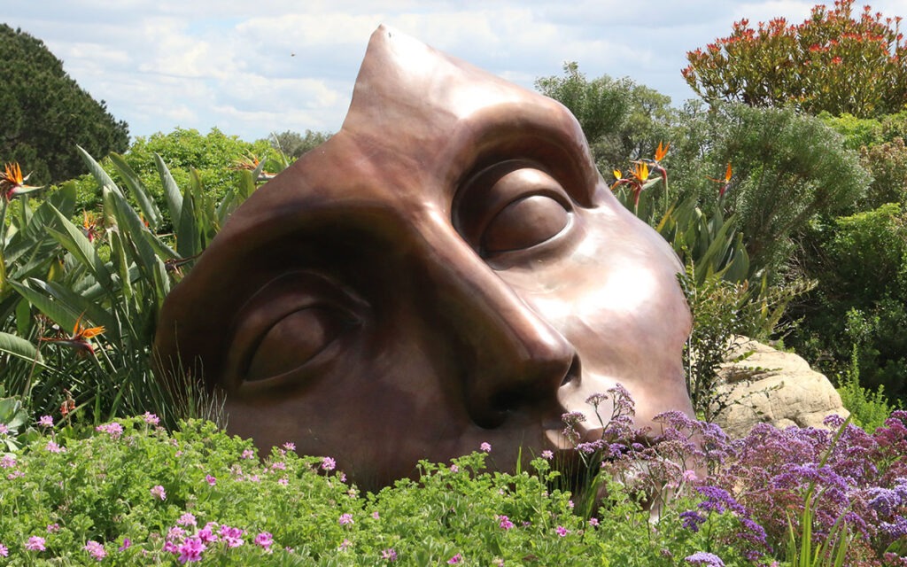 A bronze status of a large face, perched among flowering bushes in a green field.