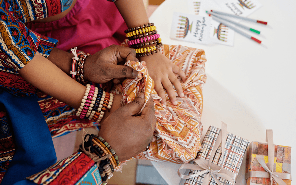 Two pairs of black hands, belonging to an adult and child, wrapping a gift in an African print cloth.