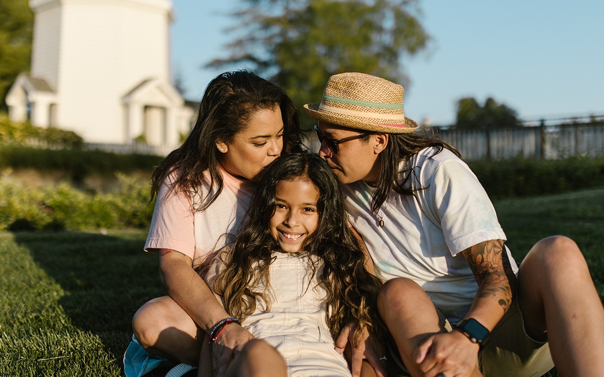 A Native American couple sitting on the grass with their daughter sitting between them. They are both kissing the child’s forehead.
