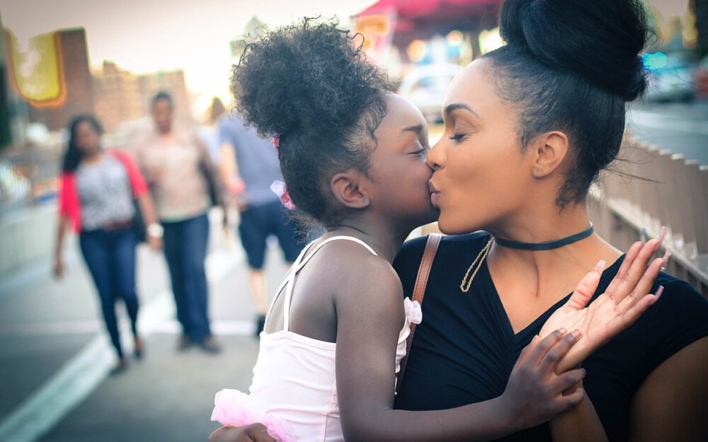A Black mother kissing her toddler daughter on the cheek and holding her in her arms.