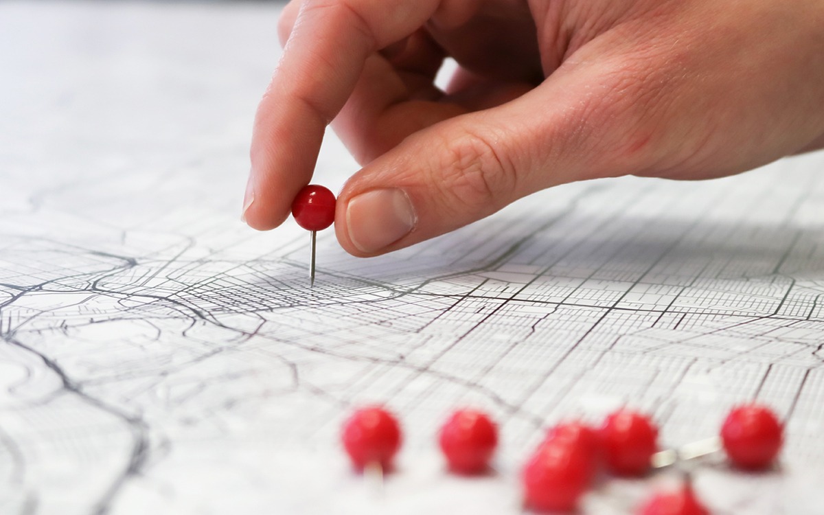  A hand placing a red push pin onto a map. There are many other red puch pins grouped together nearby.