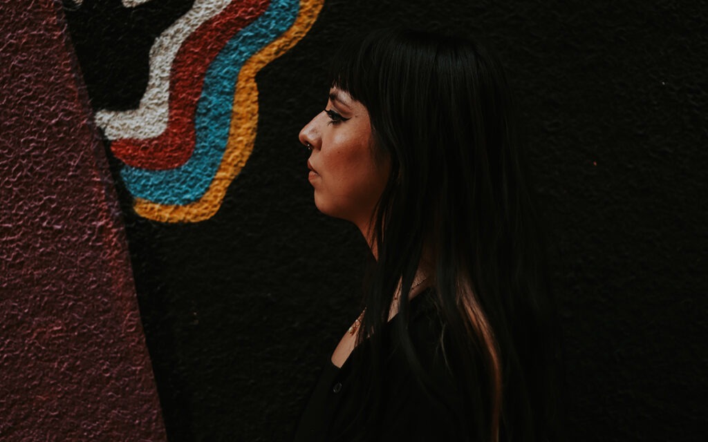 A dark-haired Latina woman with a septum piercing faces in profile to the left. She is standing in front of a concrete wall with a colorful wavy design.