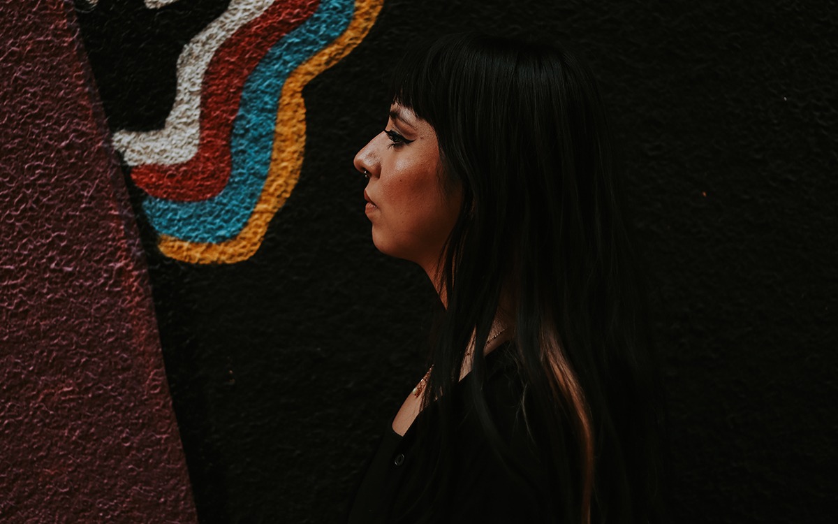 A dark-haired Latina woman with a septum piercing faces in profile to the left. She is standing in front of a concrete wall with a colorful wavy design.