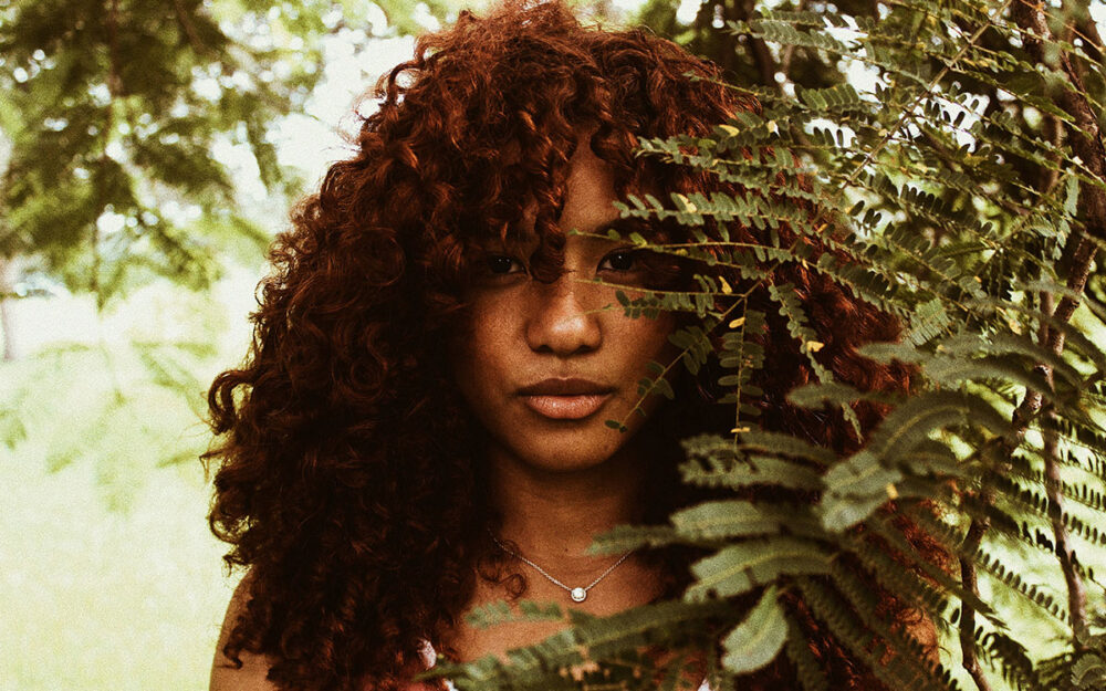 Brown woman with curly auburn hair, standing behind the branch of a tree, looking into the camera intently
