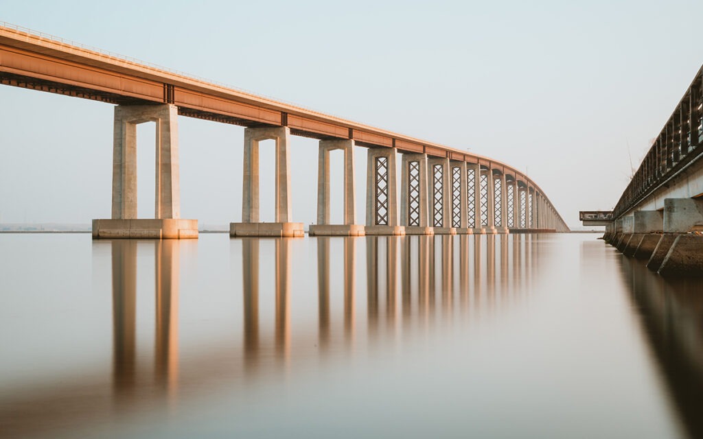 A large, concrete bridge spanning over a body of water where the bridge’s reflection is visible.